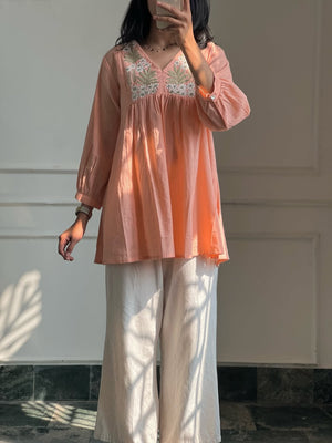 A woman wearing a peach-colored long-sleeve tunic top with white floral embroidery on the chest, paired with white wide-leg trousers, standing in front of a white paneled wall.