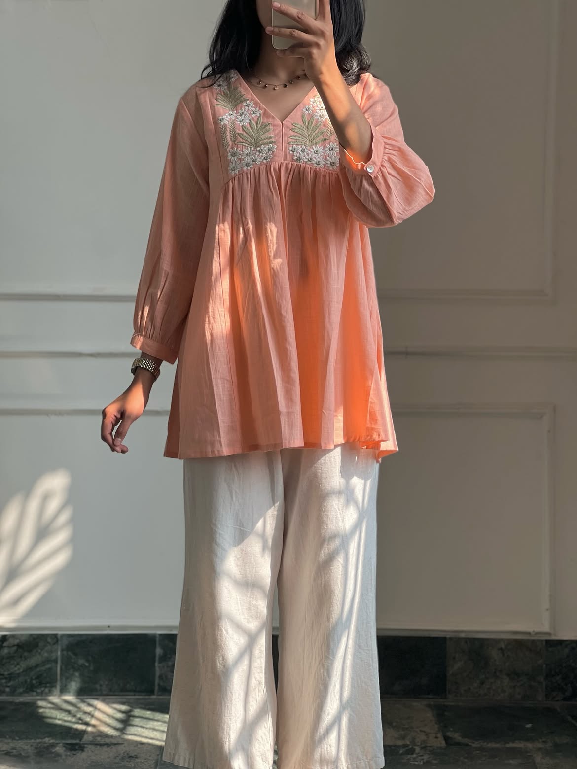 A woman wearing a peach-colored long-sleeve tunic top with white floral embroidery on the chest, paired with white wide-leg trousers, standing in front of a white paneled wall.
