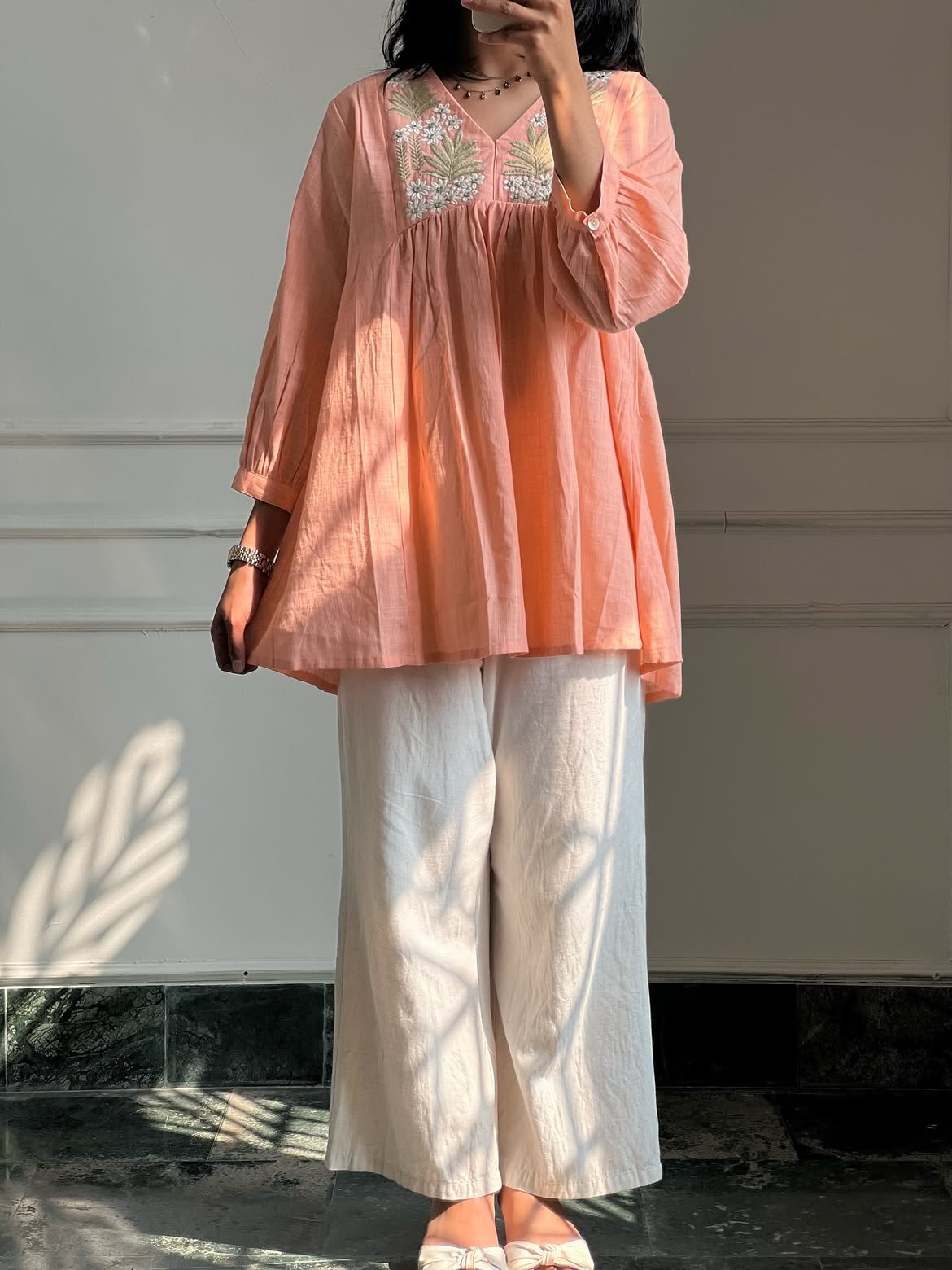 A woman wearing a peach-colored cotton tunic top with floral embroidery on the chest, paired with off-white wide-leg trousers, standing against a light-colored wall with leaf shadows.