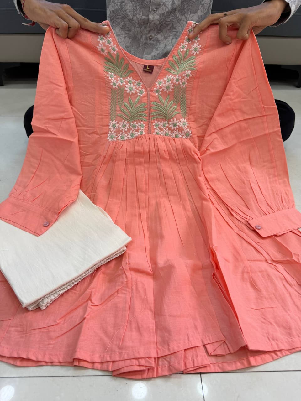 [actual-product] A coral pink embroidered tunic top being held up by hands, featuring a V-neckline with white and pink floral embroidery and green leaf motifs, paired with a white skirt.