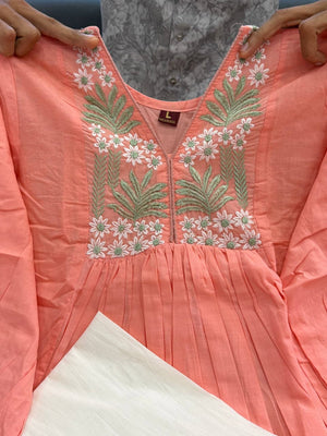 [actual-product] A coral pink embroidered tunic top being held up by hands, featuring a V-neckline with white and pink floral embroidery and green leaf motifs, paired with a white skirt.